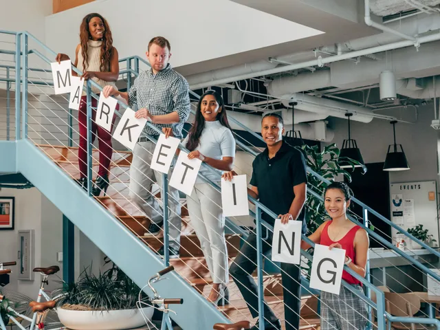 A diverse group of professionals holding 'marketing' signs on an office staircase. Event marketing agency retainer vs project fees: UK comparison